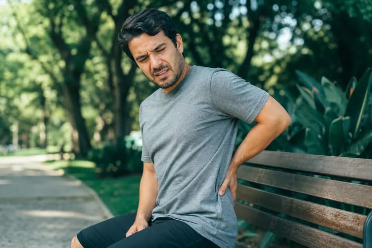 Pessoa sentada com a mão na parte inferior das costas, com expressão de dor.